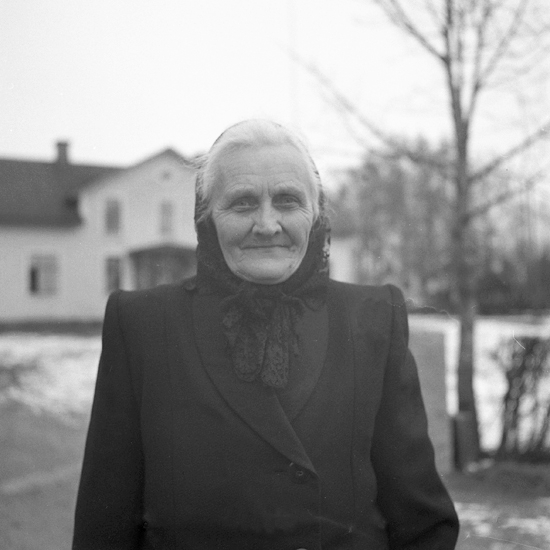 En äldre kvinna, klädd i mörk kappa och mörk s.k. kyrksjalett. I bakgrunden skymtar ett bostadshus.
Hovmantorp.