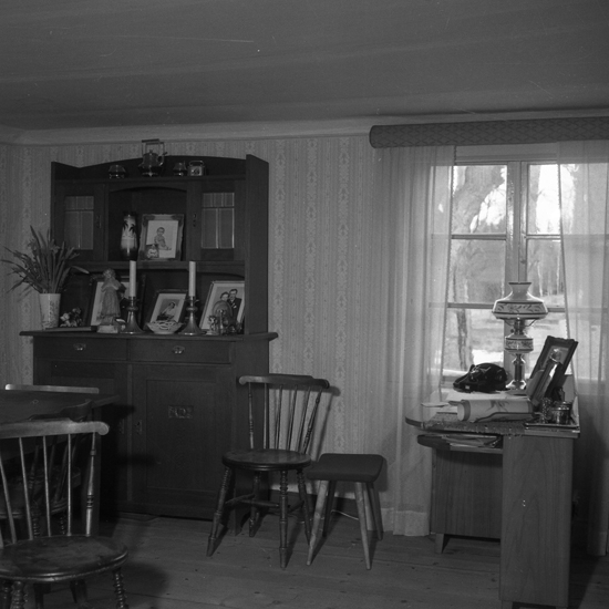 En interiör med några fotoramar, två ljusstakar, en blomvas m.m. på ett vitrinskåp.
Till höger ser man ett  skrivbord m.m.