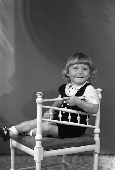 Foto av en okänd liten pojke i mörka kortbyxor och vit skjorta, som sitter på en vitmålad stol.
Helfigur. Ateljéfoto.