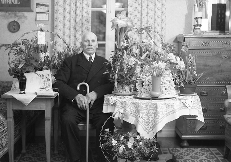 Foto av en äldre man i kostym, som sitter i ett vardagsrum omgiven av blommor och en spettekaka på ett bord.
90-årsdag. F.d. soldaten Johan Magni Lindeberg (4 februari 1865-1961), Sköldstad Södregård Germundsgård, Hjortsberga.