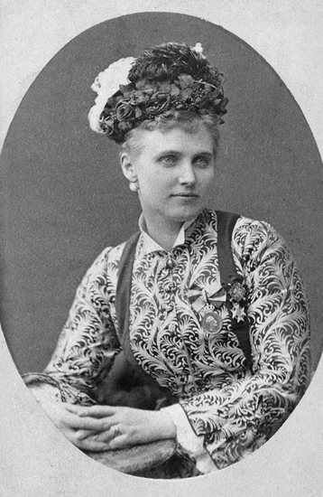Porträttfoto av Christina Nilsson. Hon bär mönstrad klänning med medaljer samt hatt och örhänge.