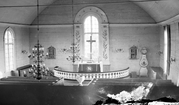 Interiör av Eringsboda kyrka sedd från läktaren. Över altargången hänger två ljuskronor. Utanför altarrundeln står dopfunten, och där innanför altarbordet med ljusstakar och ett kors. På väggen bakom hänger nummertavlorna. En klocka står till höger vid väggen.