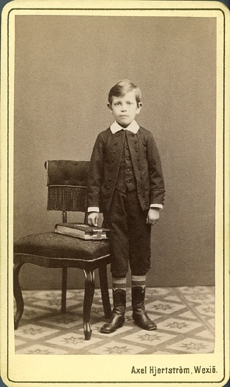 Porträtt (helfigur, en face) av en okänd pojke i knäbyxor och jacka med vit krage. Han står bredvid en stoppad, fransprydd stol, där det ligger en tjock bok (fotoalbum ?) på sitsen.
