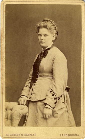 Porträtt (knäbild, halvprofil) av en ung kvinna i ljus klänning med en stor mörk rosett vid kragen. Hon lutar ena handen mot ryggstödet på en stoppad stol.