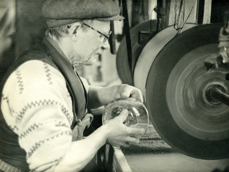 En äldre man i glasögon m.m., sitter och slipar en glasskål vid en slipmaskin.