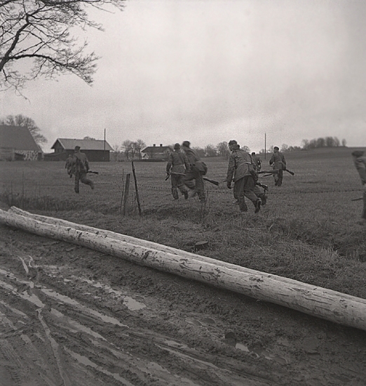 Hemvärnet. 20/4, 1941.
Några män i hemvärnsuniform skyndar iväg över en äng. I bakgrunden
syns en stenladugård m.m.