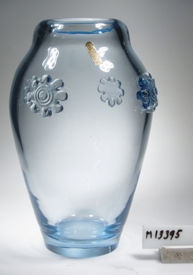 Vas.
Beskrivning: Oval vas med pålagt motiv i form av åttakantig blomma.
Färg: Blått klarglas.
Märkt med gyllene etikett. Se "Signering, märkning" ovan.
Inskrivet i huvudkatalogen 1943.
Funktion: Vas