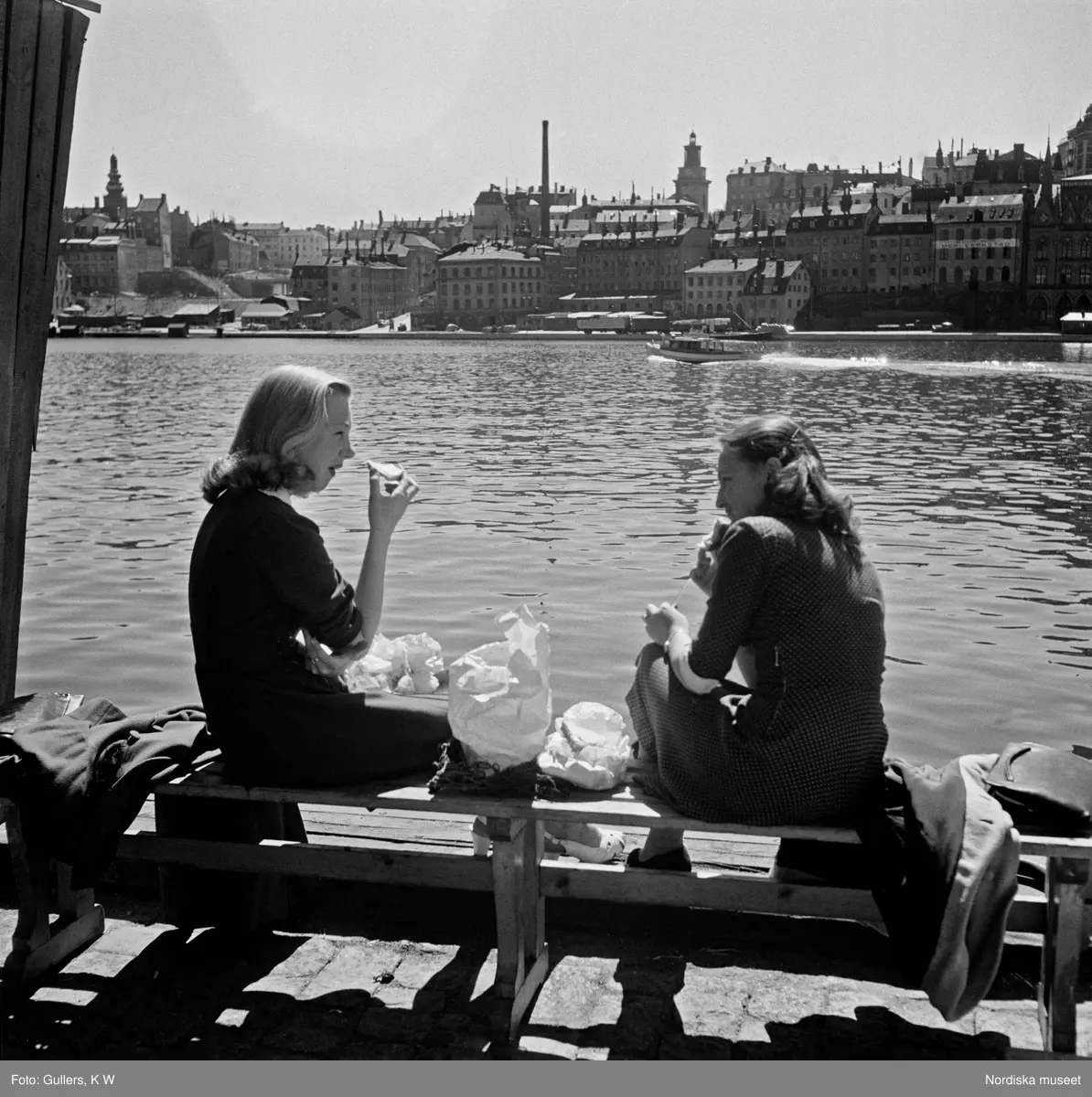 Två kvinnliga kontorister äter sin lunch vid kajen vid Södra Riddarholmshamnen, Stockholm. I bakgrunden Riddarfjärden och bebyggelse på Södermalm.