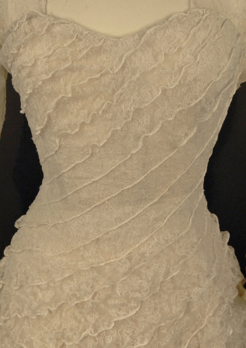 Helklipt, figursydd. Ermer og bærestykket klipt i ett. Rund hals bak med 22,5 cm lang splitt og 14 knapper med sydde trådhemper. Foran er halsutringingen rett nedover 18,5 cm fra skulderbredden ved halsen og går ned under bryststykket på kjolen. Fra bærestykket (som er foret med bobinett) er kjolen foret med tykk, blank silke. Utenpå er påsydd bobinettkapper som går på skrå fra venstre og ned mot høgre. Størsteparten av overdelen og hele skjørtet er blonden sydd på bobinettstoff uten mønster. Underskjørtet er da løst og har ei snor innsydd i folden nede for at kjolen skal føre seg penere. Glidelås i siden, hekter og sydde hemper på overdelen. Erme: Skinkeerme med 12,5 cm lang splitt ved håndleddet og 7 knapper, sydde knappehemper. 6 små innsydde legg på underarmen for bue av ermet. Skjørtet er utsvingt og har ein vidde nede på ca. 668 cm. Vidden på underskjørtet 470 cm. Kjolen er 19 cm lengre bak enn foran.