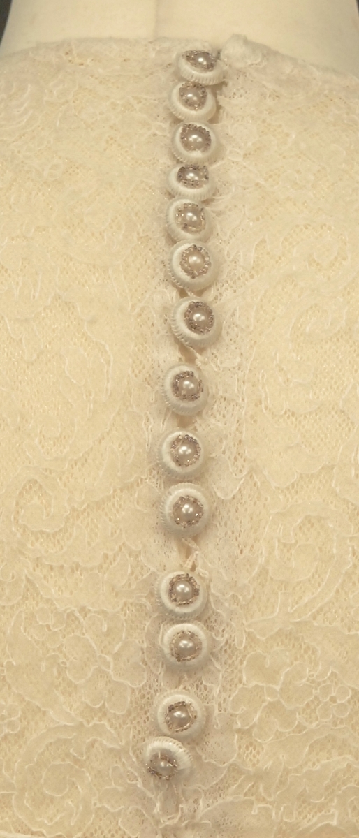 Helklipt, figursydd. Ermer og bærestykket klipt i ett. Rund hals bak med 22,5 cm lang splitt og 14 knapper med sydde trådhemper. Foran er halsutringingen rett nedover 18,5 cm fra skulderbredden ved halsen og går ned under bryststykket på kjolen. Fra bærestykket (som er foret med bobinett) er kjolen foret med tykk, blank silke. Utenpå er påsydd bobinettkapper som går på skrå fra venstre og ned mot høgre. Størsteparten av overdelen og hele skjørtet er blonden sydd på bobinettstoff uten mønster. Underskjørtet er da løst og har ei snor innsydd i folden nede for at kjolen skal føre seg penere. Glidelås i siden, hekter og sydde hemper på overdelen. Erme: Skinkeerme med 12,5 cm lang splitt ved håndleddet og 7 knapper, sydde knappehemper. 6 små innsydde legg på underarmen for bue av ermet. Skjørtet er utsvingt og har ein vidde nede på ca. 668 cm. Vidden på underskjørtet 470 cm. Kjolen er 19 cm lengre bak enn foran.