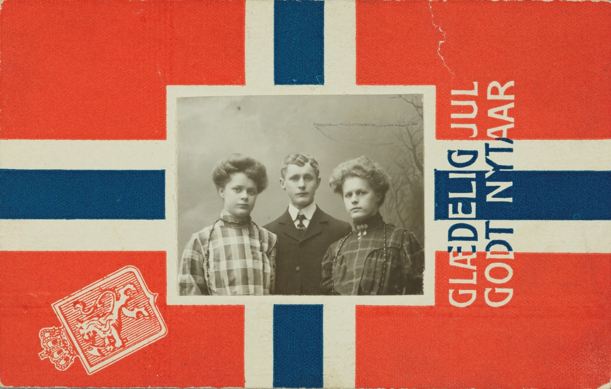 Jule- og nyttårskort. Portrett av tre ungdommer, to jenter og en gutt, på en bakgrunn med det norske flagget med riksvåpenet. Datert 1905.