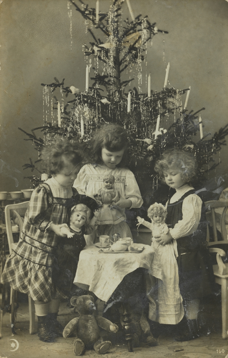 Julekort. Jule-og nyttårshilsen. Fotografi. Tre pyntede piker med dukker, bamse og koppestell foran et juletre med stearinlys.