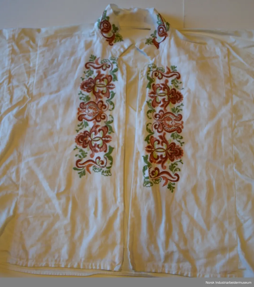 Plattsøm på kraven, mansjettene og bringa. Skjorten er åpen foran og helt ned.