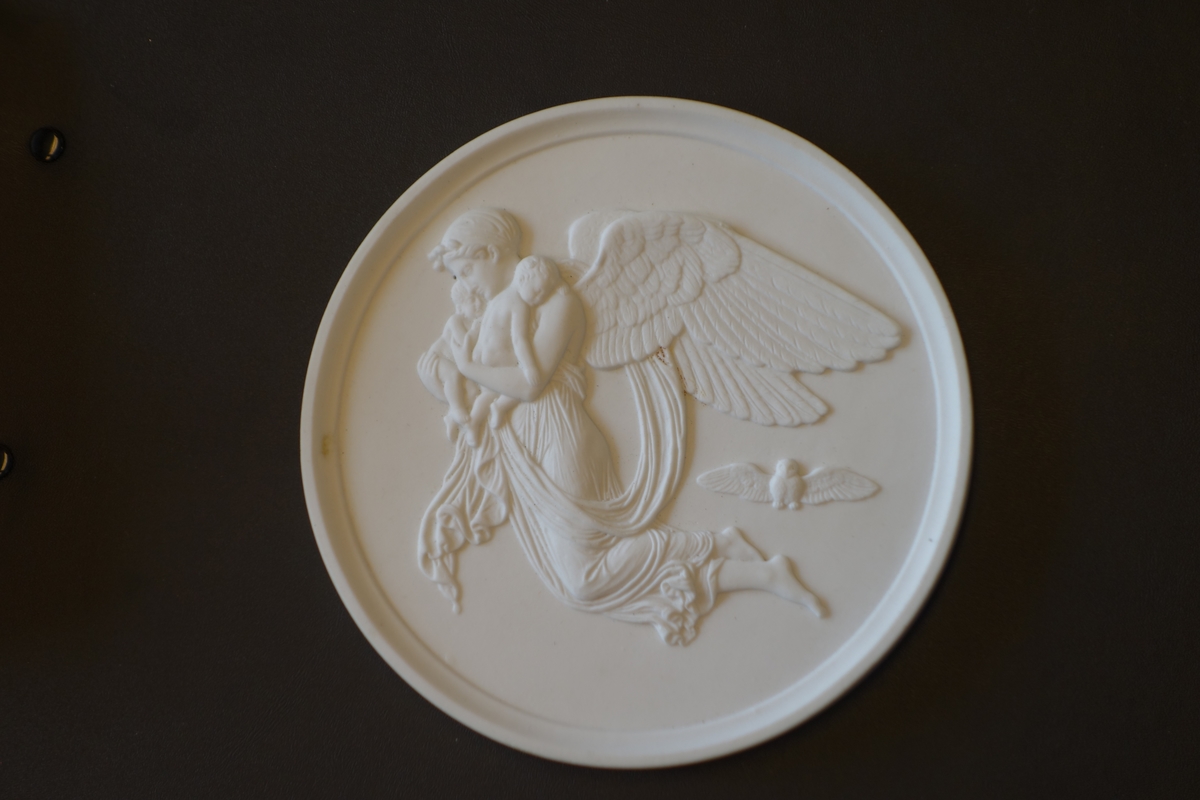 Rundt porselensrelieff med profilert kant. Motivet i relieffet er en bevinget kvinnefigur som svever med to barn i armene, mens en ugle flyr under vingene hennes.