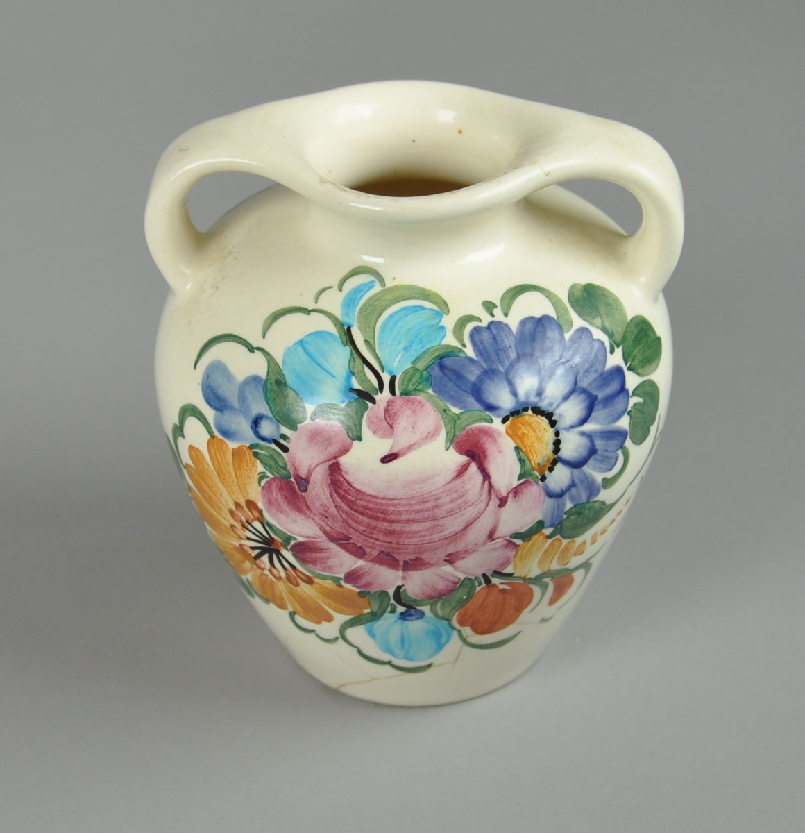 Vase av glassert keramikk, med to håndtak og dekormaling. Motiv på maling av blomsterkrans. Vasen har en utstående form. Vasen er noe oppsprukket ved bunnen.