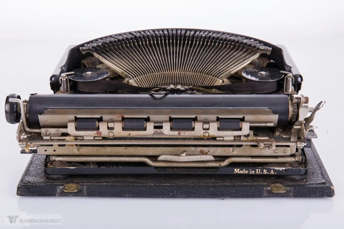 Mekanisk skrivemaskin i metall med 46 tangenter. Metallnøklene ligger åpent oppe på maskinen i en halvsirkel med fargebåndruller på hver side. Karosseriet er avrundet og skrudd fast i en treplate som festets til maskinens transportkasse som har låsemekanisme og bærehåndtak. Nøkkel følger med.