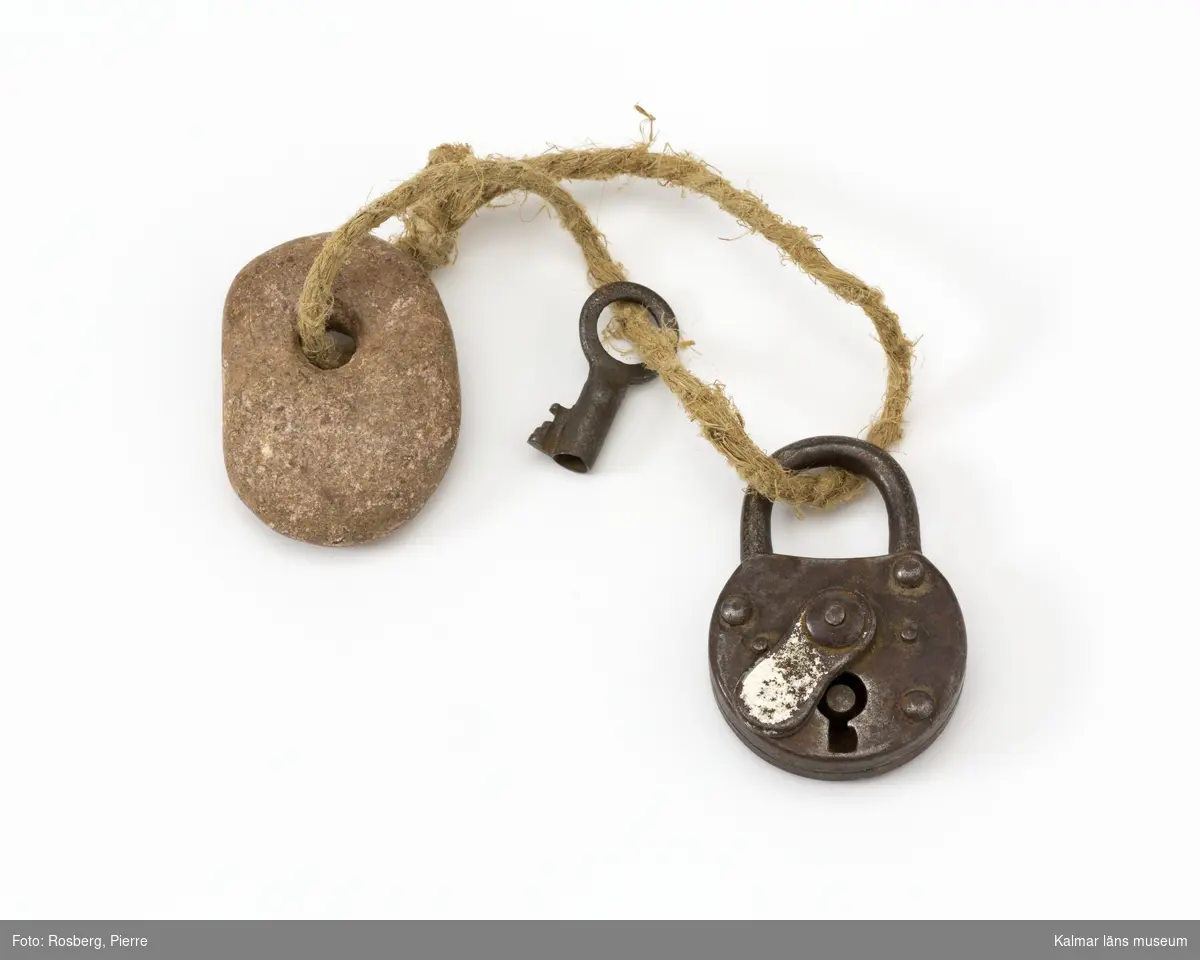 KLM 28979 Hänglås, av metall, sten och snöre. Hänglås med bricka framför låset, nyckel och nyckelbricka av sten. Allt hålls samman av ett slitet snöre.