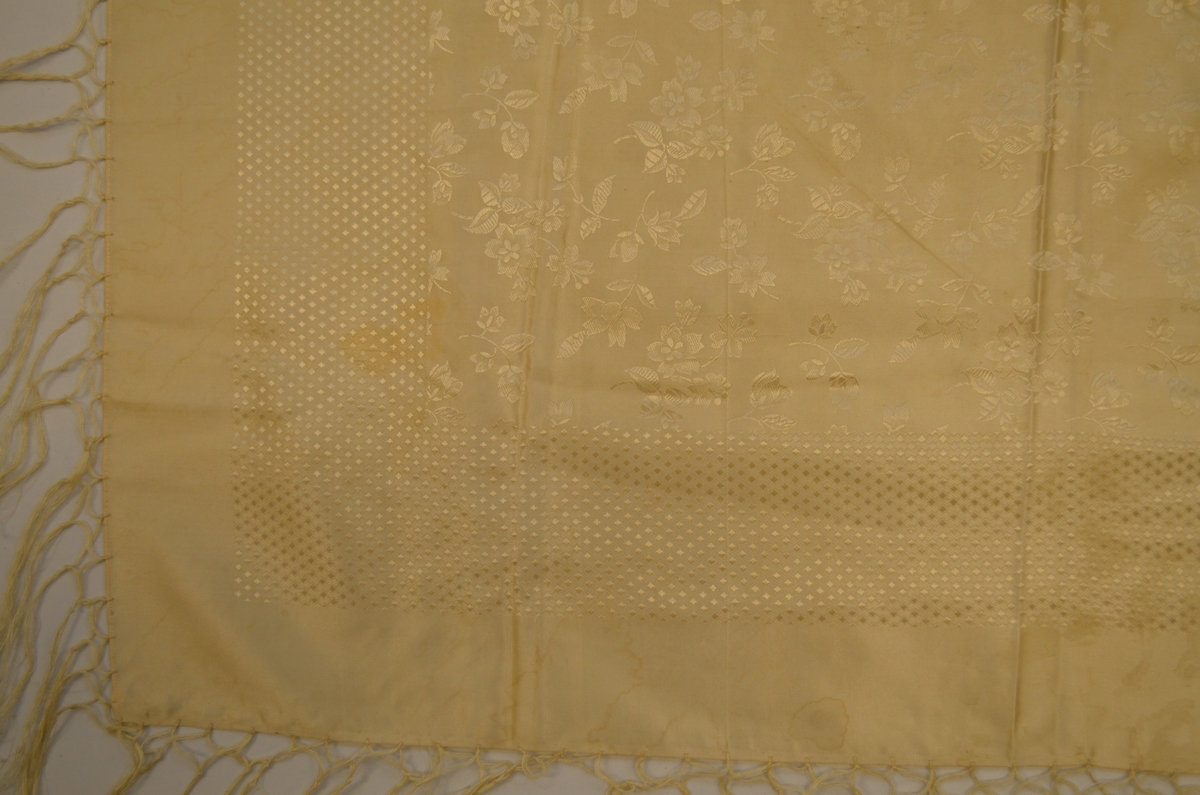 Tørkle i beige silkedamask med lysebrune rosebordar langst kantane. Påsette frynser i beige 10,5 cm lange.
