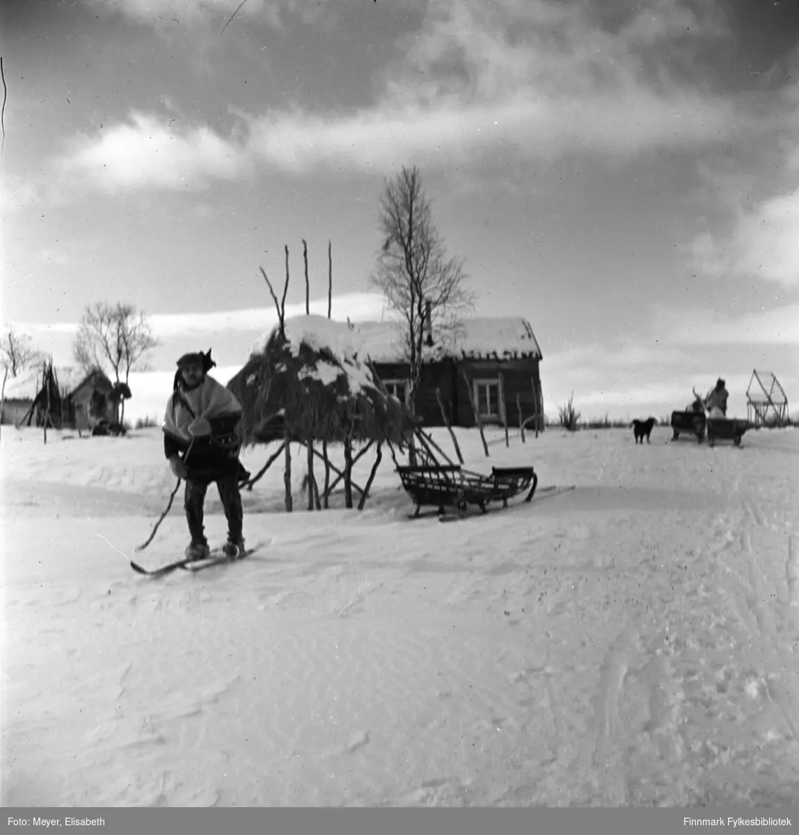 Vinterhus til reindriftssamer. Same på ski med bjørkekjepp som stav. I bakgrunnen sees en slede, et vinterlager og flere hus. Til høyre i bildet sees en same med slede og en hund ved siden av. Bildet er tatt i 1939-40.