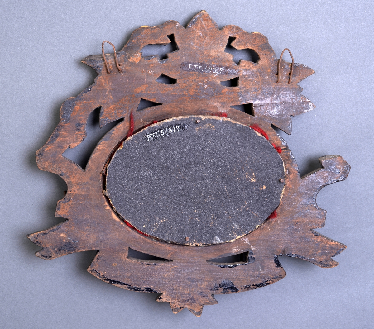 En knaggrekke som består av et brodert bilde som er rammet inn. Bakgrunnen på bildet er brodert med rødt garn (ullgarn?). På midten er det brodert en liggende hund i glassperler i ulike nyanser av brunt og grått samt hvite glassperler. Broderiet er rammet inn i en ramme av tre. Rammen rundt det broderte bildet er ovalt med enkel profilering. Rundt denne rammen er det utskjæringer. På oversiden er det relativt store blader som er skåret ut. På undersiden er det et slags banner som bølger seg. I denne er det festet fem enkle metallkroker til å henge ting på. En del av utskjæringene mangler på vesntre side. Rammen har nok vært lakkert. På baksiden er det festet to enkle hemper laget av ståltråd til oppheng. Det er skrudd på ny plate på baksiden.