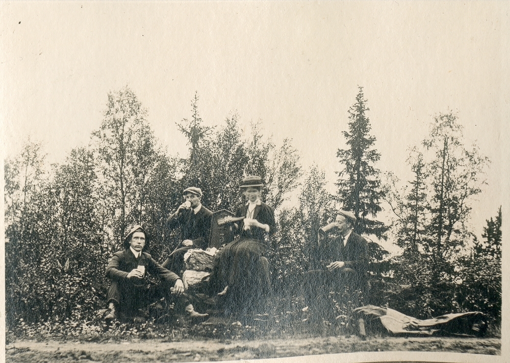 Gruppe. Utendørs. Rast i naturen. Tre menn og en kvinne sitter ute i skogen, drikker og spiser.