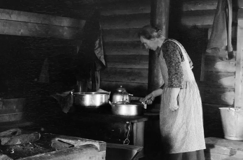 Skogskokke som står ved ovnen og lager mat i ei gammel koie