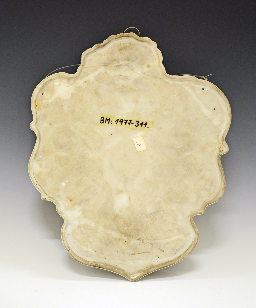 Fra protokollen: 
Porselensplatte:  "Den norske Nordpols-expedition 1893-1896", laget på Porsgrunds Porselænsfabrik. Platten har hvit bunn og er formet som et løvblad, kantet i blått og gull. Øverst det norske riksvåpen, ellers bilder av alle polfarerne, med Fritjof Nansen i midten. Rundt dette bildet laurbærkrans i grønt med sløyfe i de norske farger. I alt 13 personer. Nederst en del av en globus med tegning av polarskuta "Fram" på Nordpolen.