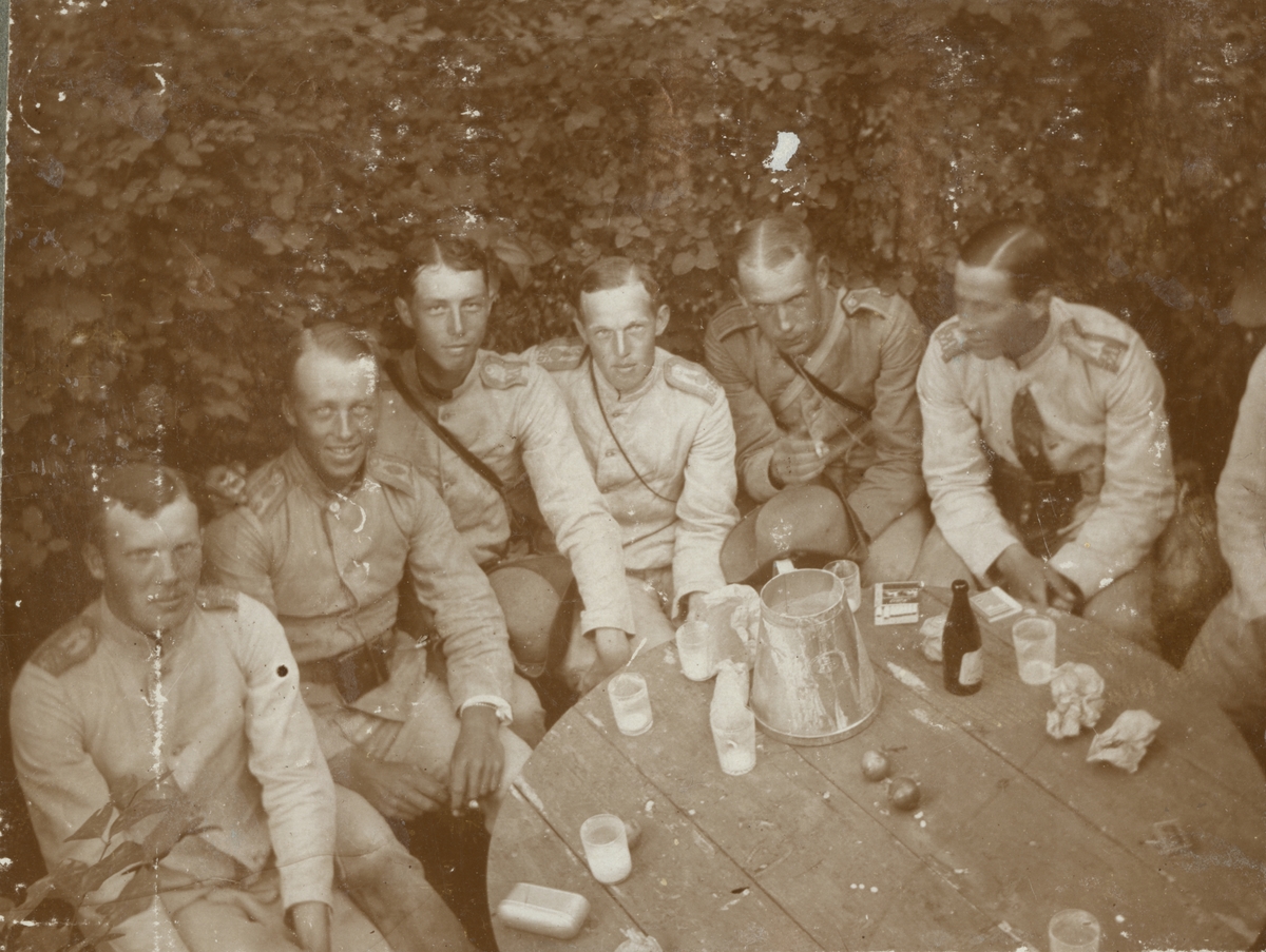 En grupp soldater kring ett bord i trädgården. På bordet finns bland annat en mjölkanna, ett antal glas och två äpplen.