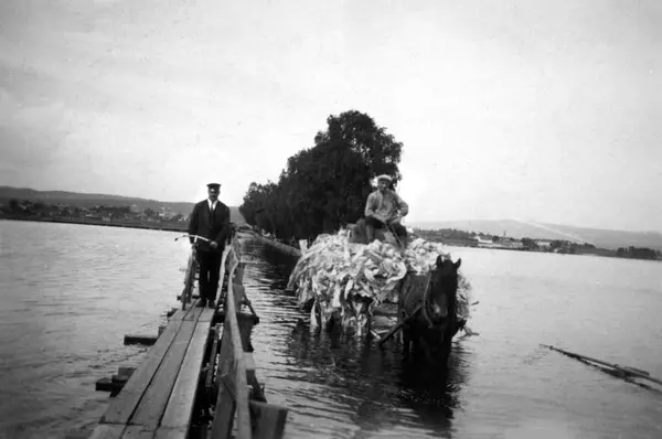 Svart-hvitt foto fra 1927. Gangbru med mann og hestetransport med stort lass som kjører på ordinær bru i flomvann.. Foto/Photo