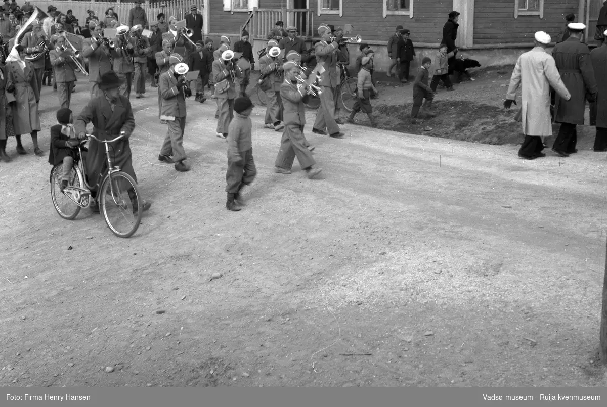 Vadsø 17. mai 1951. Motivet er fra Nyborgveien. Guttekorps spiller.  Gjerdet øverst til venstre er Esbensengården på Bakken, og huset ved siden av er Vartigården.  En mann triller en sykkel med et barn bak på gabasjebrettet.