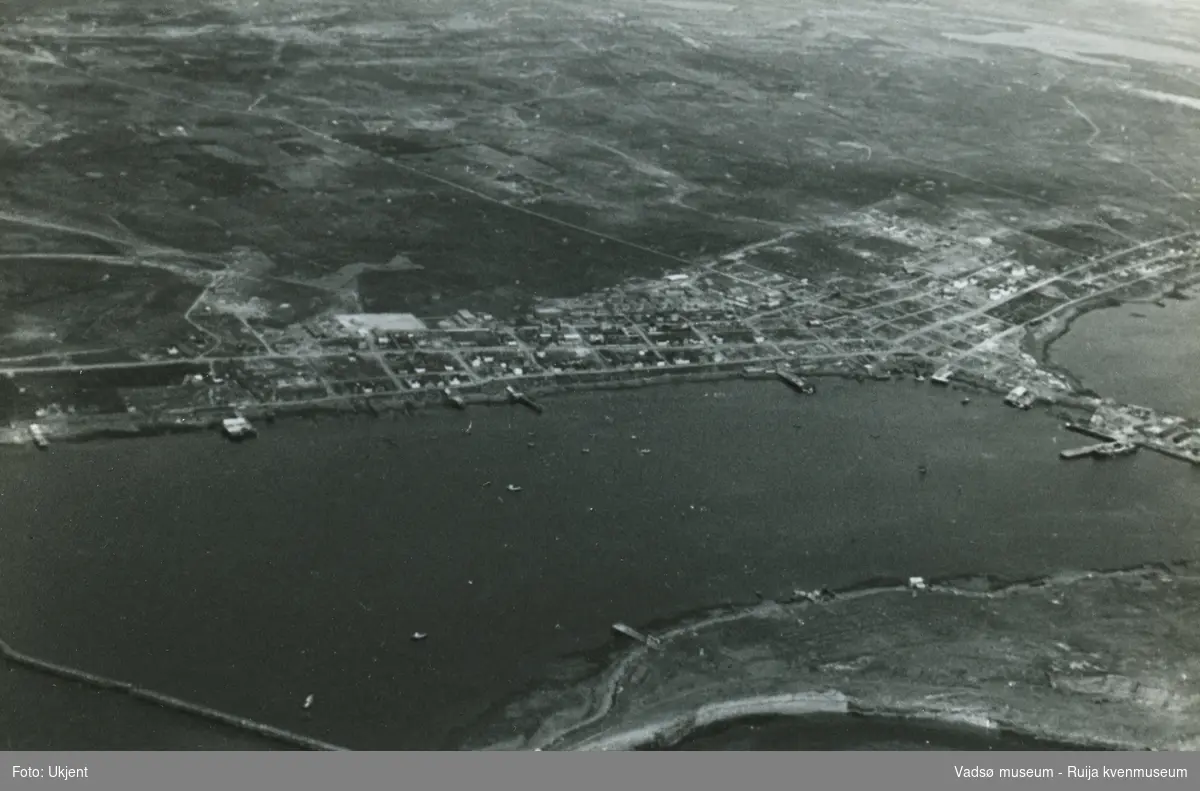 Flyfoto av Vadsø etter 2. verdenskrig i 1945. Bildet viser Havna med Indrebyen, Sentrum, Ørtangen, deler av Ytrebyen og deler av Vadsøya. Vi skimter båter ute på havna. Fra Øya ser vi moloen og skimter en del bebyggelse og kaier ved sjøkanten.  Fra midtbyen ser vi Fossevegen opp mot utmarka.