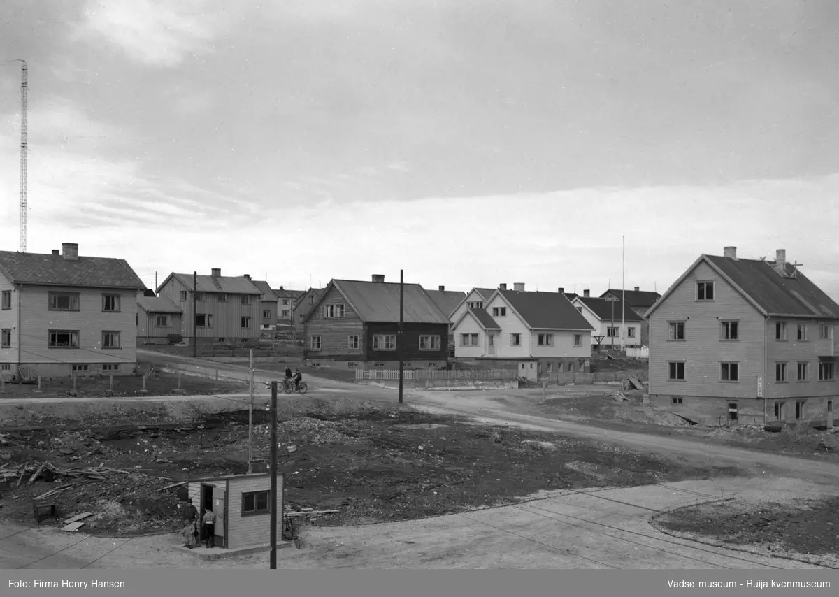 Gjenreisningsbeyggelsen i Vadsø i Midtbyen. Vi ser typiske kataloghus fra Den Norske Stats Husbank. Et næringsbygg med frisør, og en ubebygget tomt. Bildet er tatt mot nordøst.