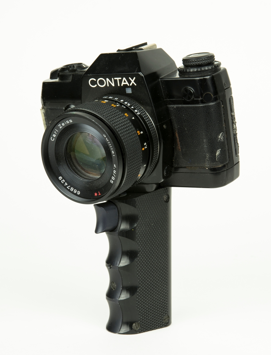 Handkamera HK 101 MT. På handkameran finns ett objektiv. Under kameran finns ett längre handtag varpå det finns 2 knappar för utlösning av film.