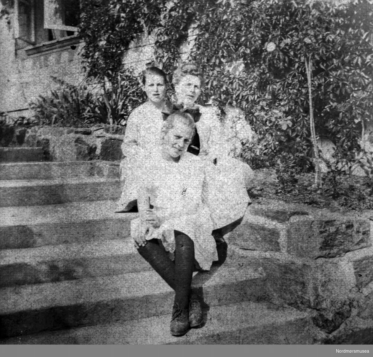 Foto av tre kvinner som sitter på en trapp. Trolig fra familien Williams i Kristiansund, og sannsynligvis fotografert omkring 1918-1920. Fra et fotoalbum tilhørende Ellinor Williams-Phakdikun (1905-1963). Fra Nordmøre museums fotosamlinger.