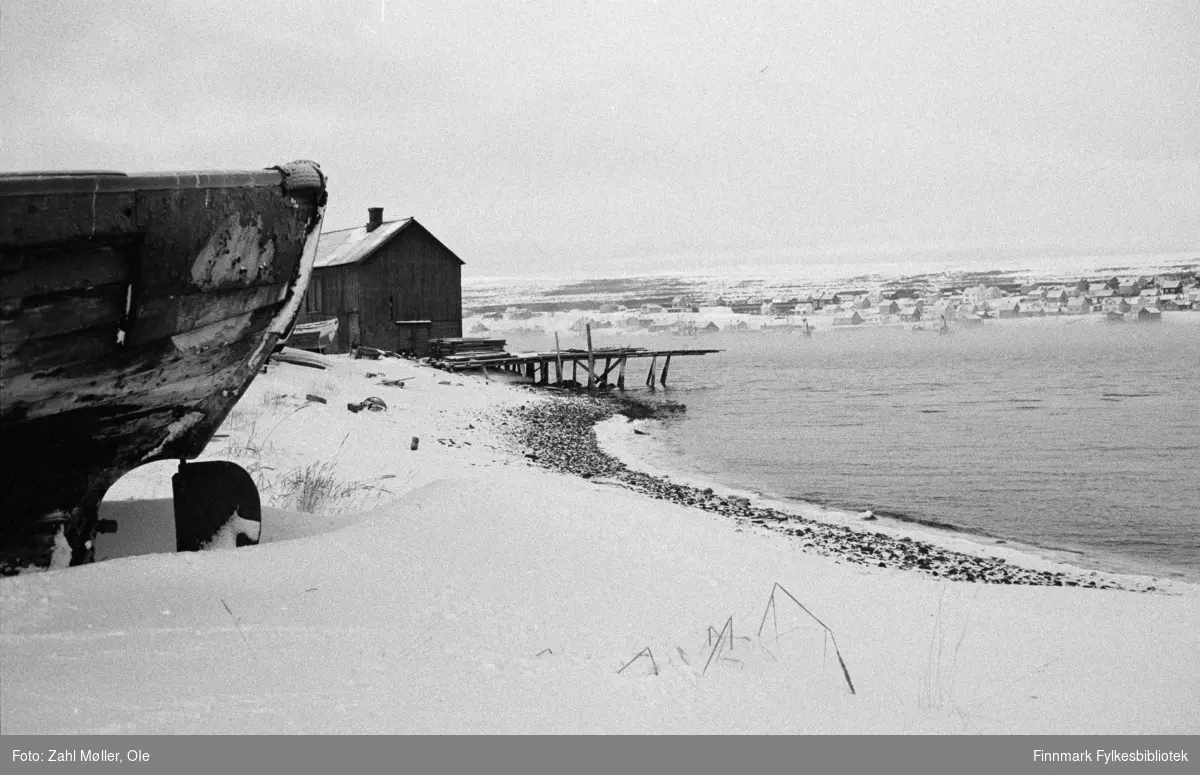 Vadsø 1968-69. Dette vinterbildet er tatt ute på Vadsøya fra området ved Sildoljefabrikken/Vadsø Havn. I venstre forkant ser vi skroget på en båt som ligger på land. I bakgrunnen et gammelt kaianlegg med en kai som ser ut til å gå i oppløsning. I bakgrunnen sees Vadsøs bebyggelse. Det er skodde eller sterk vind på fjorden.
