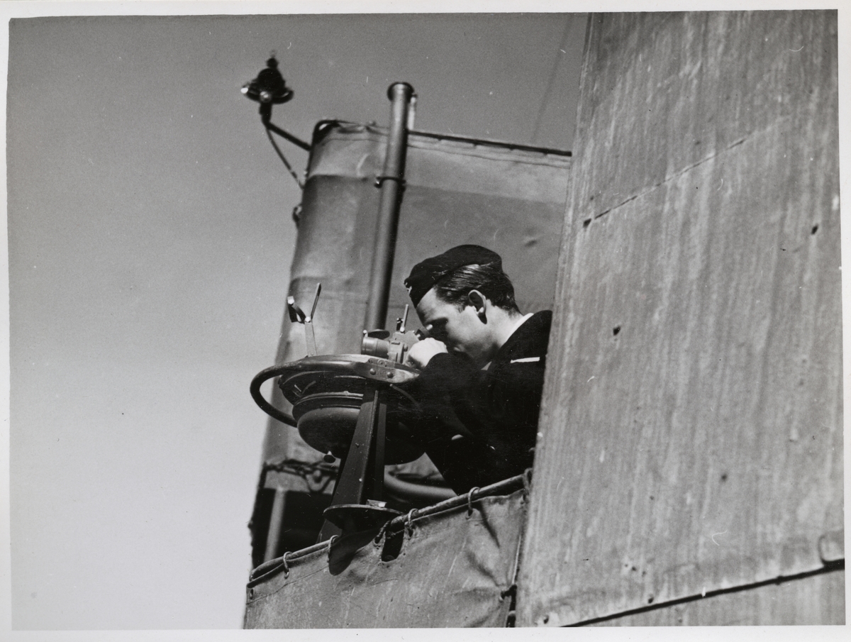 En sjöman tittar genom en kikarsikte på bryggan av en örlogsfartyg.