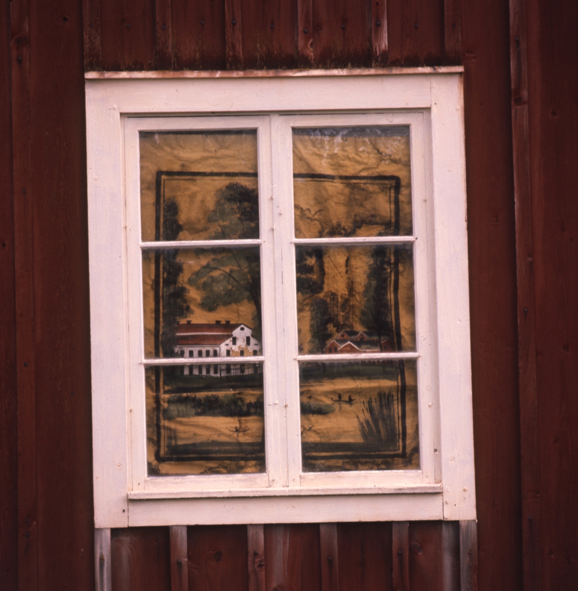 Exteriör med spröjsat fönster på röd trävägg. Innanför fönstret ser man en rullgardin med mönster föreställande ett landskap med byggnader.
