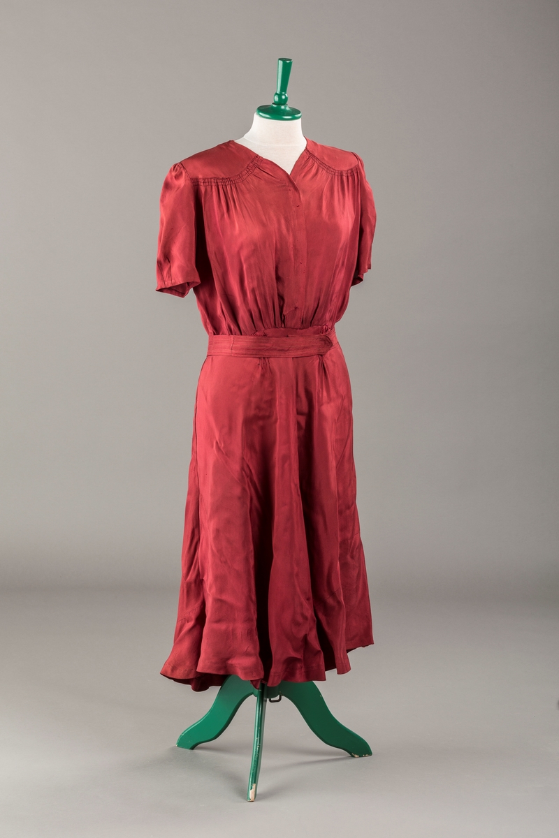 Form: Glatt, klokkeformet skjørt. Forstykkene rynket til buede bærestykker foran. Overdel rynket til skjørt. Korte ermer. Lukket med trykknapper. Belte fastsydd.