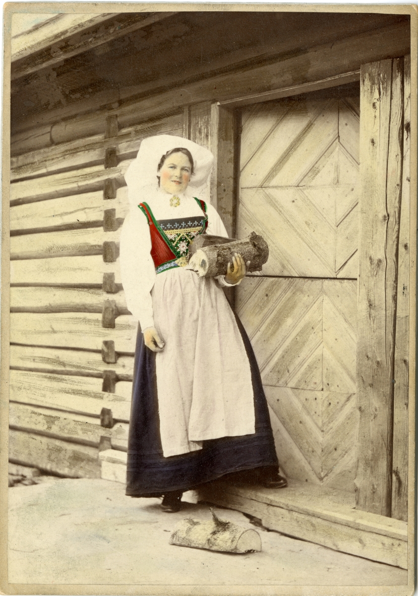 Kolorert fotografi av kvinne med drakt, bærer ved står foran dør. Hardanger.