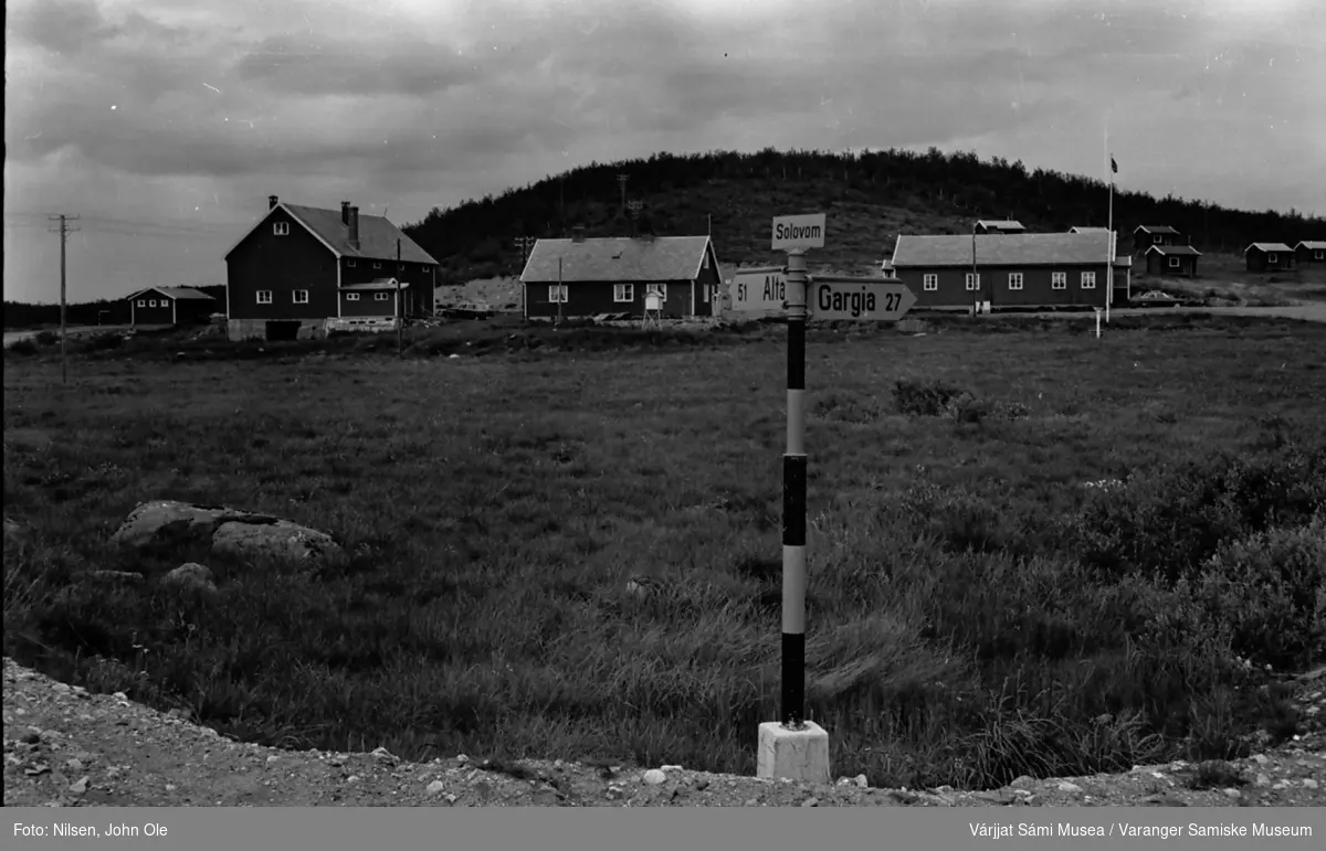Solovom fjellstue. Vegskilt med teksten:  "Solovom. Avstand til Alta 51 km og til Gargia 27 km." i forgrunnen. Fjellstua og flere hytter i bakgrunnen. Juli 1967.
