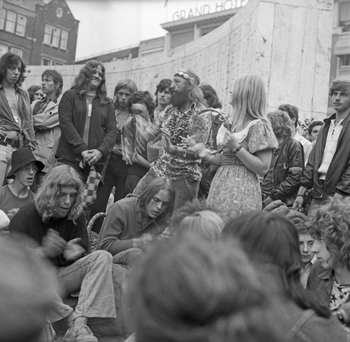 En gjeng som er samlet og spiller og synger på en åpen plass i sentrum av en by. Muligens Amsterdam.