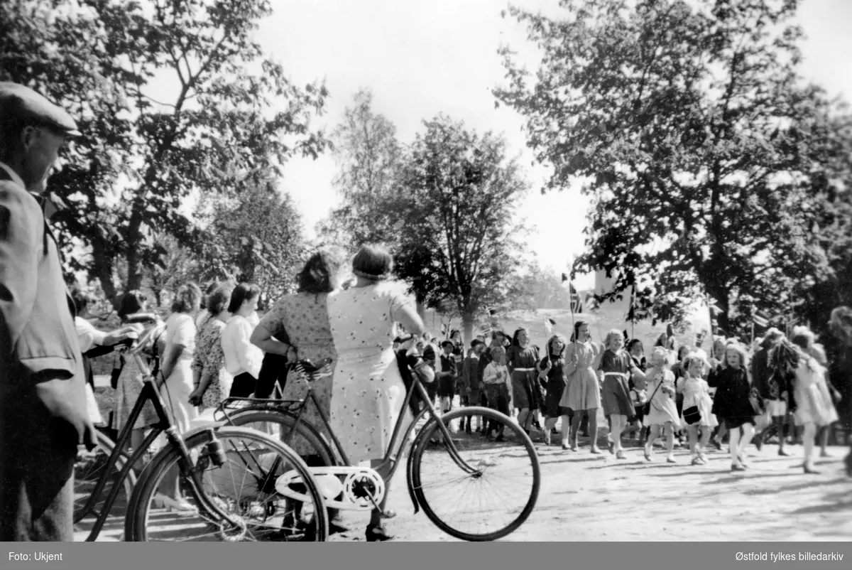 17. maitog med tilskuere med sykkel i Skiptvet sentrum, Karlsrud krets, ca. 1955.
Personen til venstre er Kristian Vister f. 1892. Sykkelen var framkomstmiddel for alle.