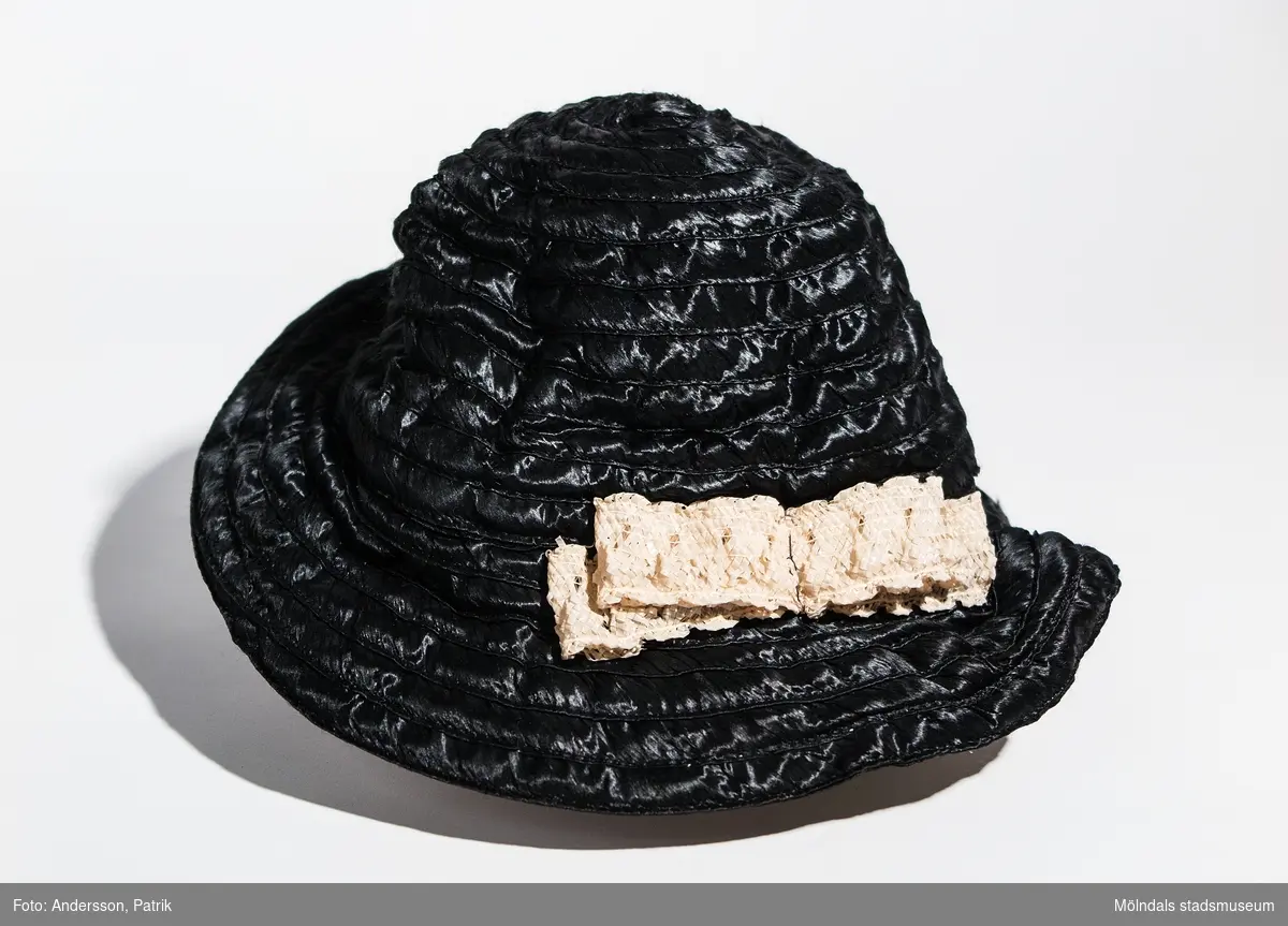 Damhatt inköpt i Eskilstuna, troligtvis på 1930-talet.
Hatten är svart. Den har vit spets som detalj fastsydd på vänster sida av hatten och har ett svart gummiband som hakband.
I hatten finns en etikett fastsydd med texten: 
TEL. 3199 Elsie ESKILSTUNA KÖPMANGAT.52

Enligt uppgift från givaren är hatten inköpt på 1920-talet, men eftersom butiken öppnade 1934 är hatten troligen inköpt på 30-talet.