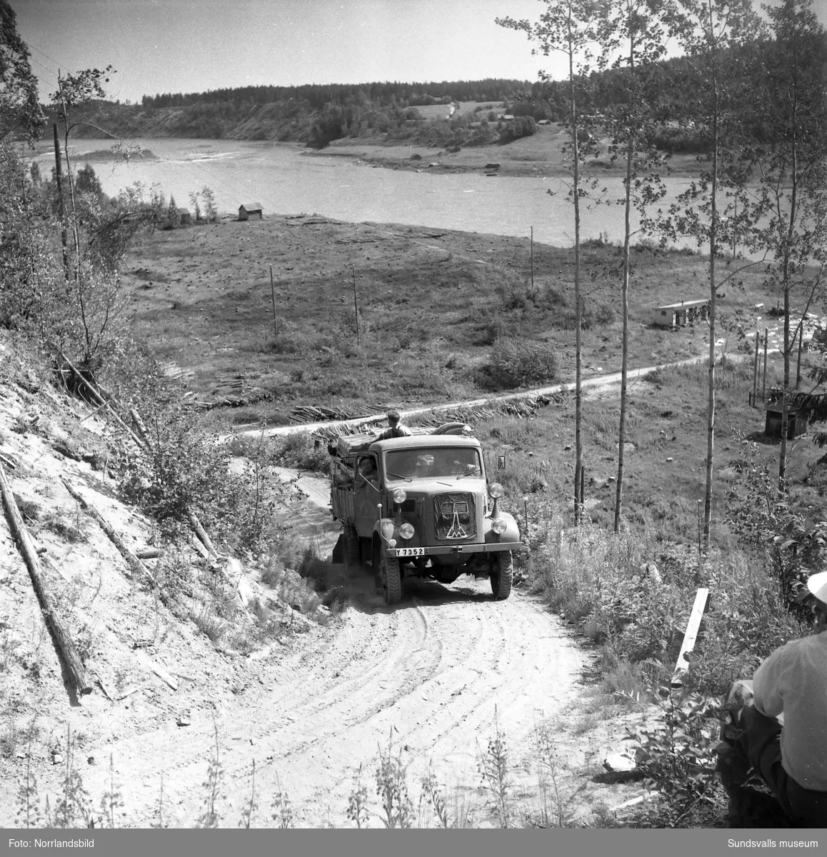 En lastbil körs uppför en brant grusväg invid en älv. Näslund & Burestad.