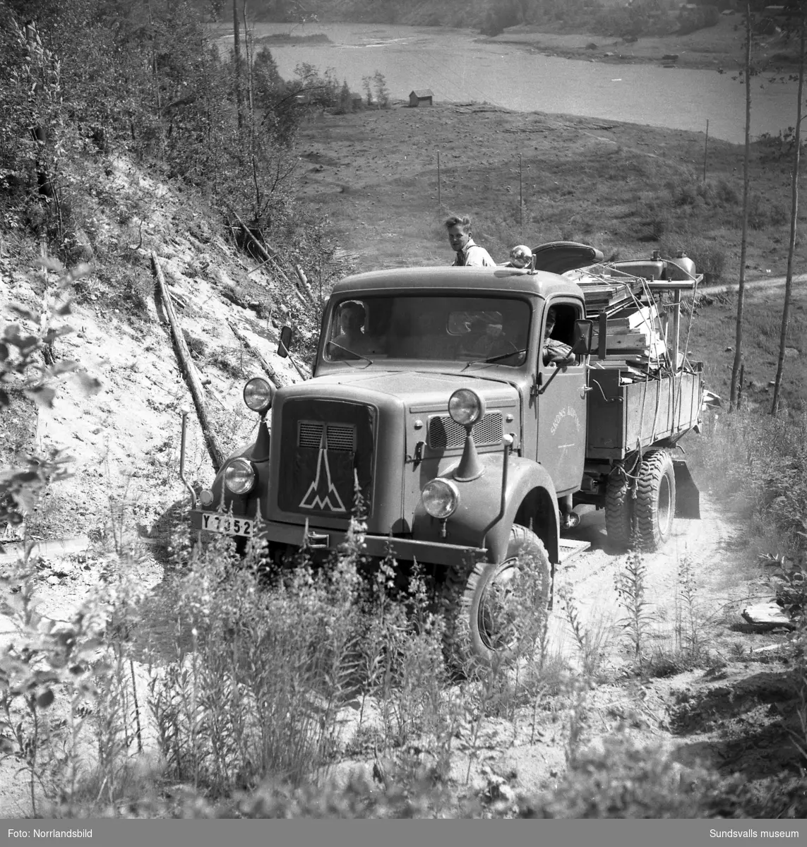 En lastbil körs uppför en brant grusväg invid en älv. Näslund & Burestad.