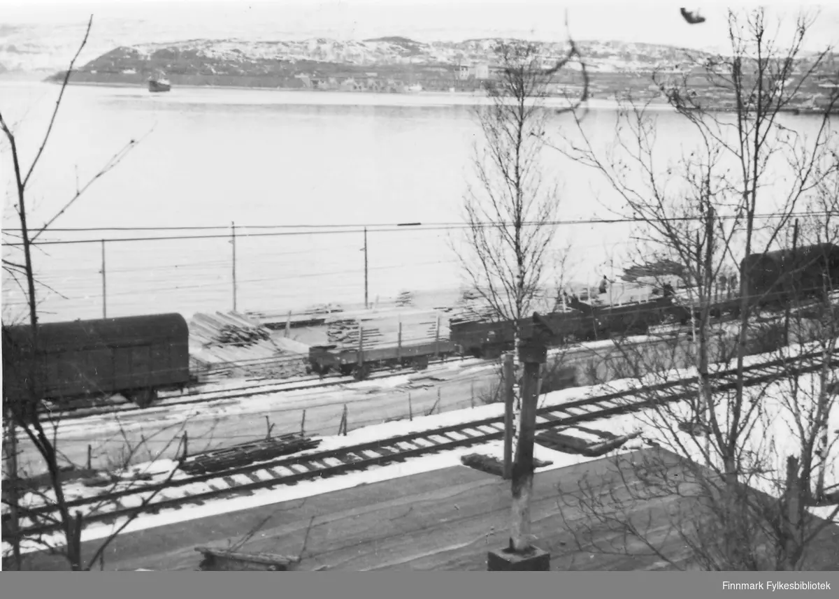 Før og etter 1944, Nordland, Troms. Utsikt mot kaia med lager av trematerialer. Jernbanen sees i forgrunn.Havet ligger rolig i bakgrunn. På andre siden av sjøen har man utsiktmot L.K.A.B anlegg. Sneen ligger ennå på marken.