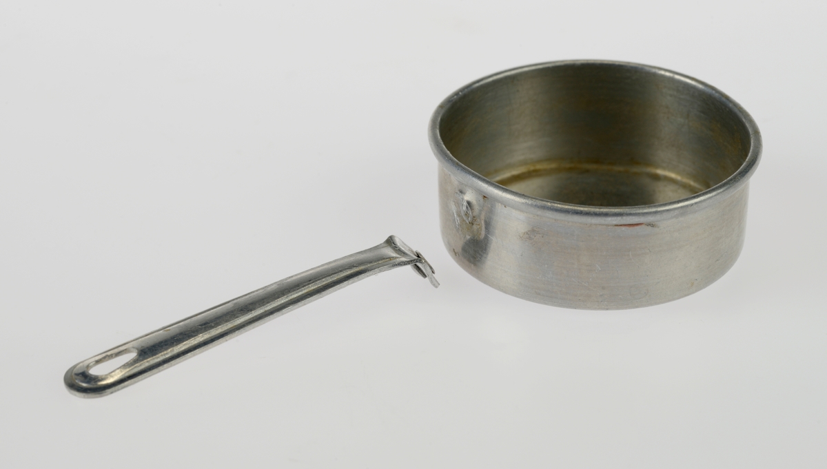 En liten komfyr med tre kasseroller og en steikepanne. Gjenstandene er undernummerert 01-05.
FTT.54454.01: En svartlakkert lekekomfyr som er firkantet med avrundede hjørner. Den er trolig laget av jernblikk og blikk. På kokeflaten er det tre hull for å plassere kasserollene på. Bakerst til venstre er det en 8cm høy pipe. I front er det en sølvfarget blikkdør som er festet med hengsler. På døren er det innskrift og et lite messingfarget og rundt håndtak. Komfyren har buede bein.
FTT.54454.02: En høy kasserolle laget av blikk. I hver side er det loddet på håndtak. 
FTT.54454.03: En lav kasserolle laget av blikk. I hver side er det loddet på håndtak.
FTT.54454.04: En lav kasserolle laget av blikk. Det har vært festet et langt håndtak i den ene siden, men det er nå løs.
FTT.54454.05: En steikepanne laget aav blikk. Håndtaket mangler.