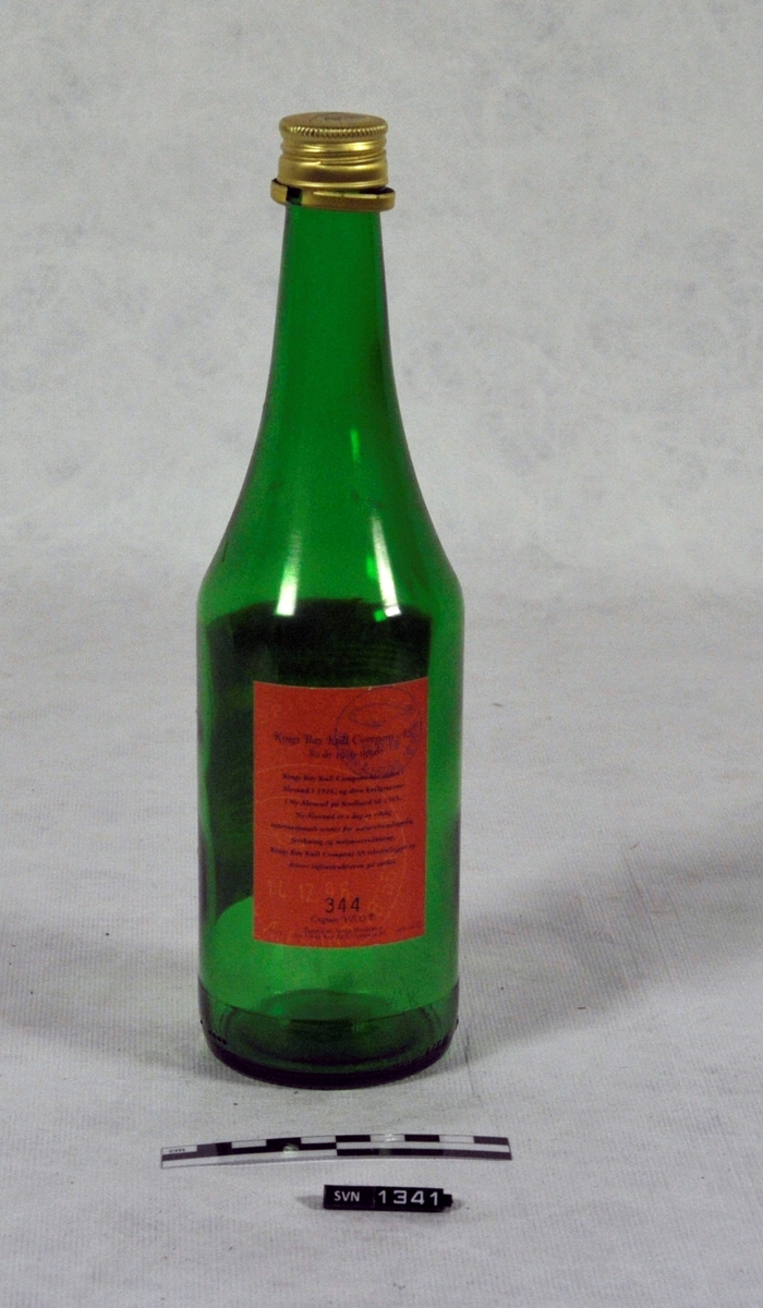 Trekasse med treflis og en fjær.
Kassen inneholder to flasker ( B og C)
Flaskene er jubileumsflasker, en fra Kings Bay Kull Company og en fra Store Norske Spitsbergen Kullkompani.
Kassen er delvis åpen på en side.
Det er en skillevegg mellom de to flaskene.
Flaskene er tomme
Flasken B er av grønt glass med rød/rosa etikett. Flasken er en jubileumsflaske for Store Norske Spitsbergen Kullkompani Flasken har skrukork

Flasken C er av grønt glass med hvit etikett med malt bildetrykk. Flasken er jubileumsflaske for Kings Bay Kull Company. Flasken har ikke kork.