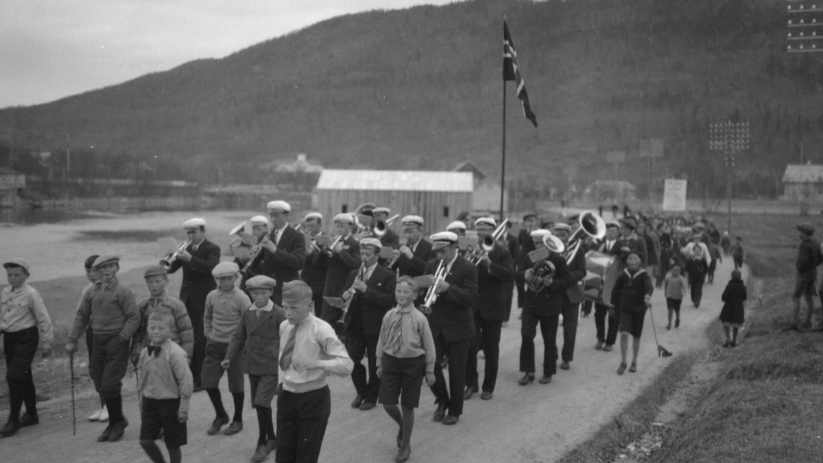 17 Mai tog i skjervgata. Hornmusikk. Båtbyggeriet til Mjaavatn med lyst tak. Johan Bech Olsen i første rekke i korpset, nr. 3 fra venstre.
