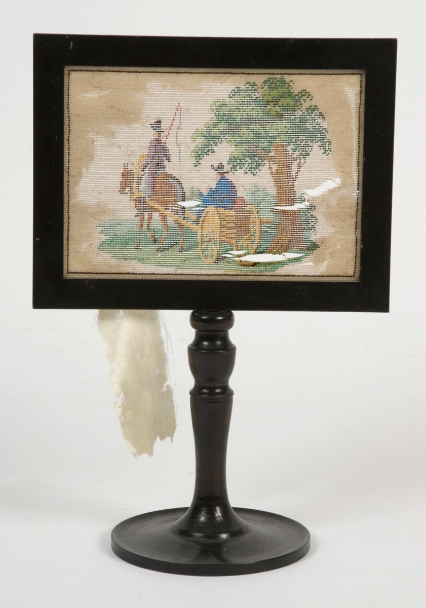 Perlebroderi spent opp på i treramme stående på en dreid trefot.  Motiv ridende mann med vogn med mann og et tre.